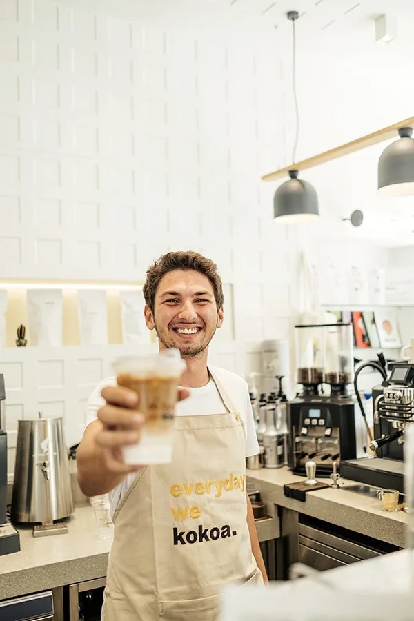 A barista in an 'everyday we kokoa.' apron holding out an iced coffee.