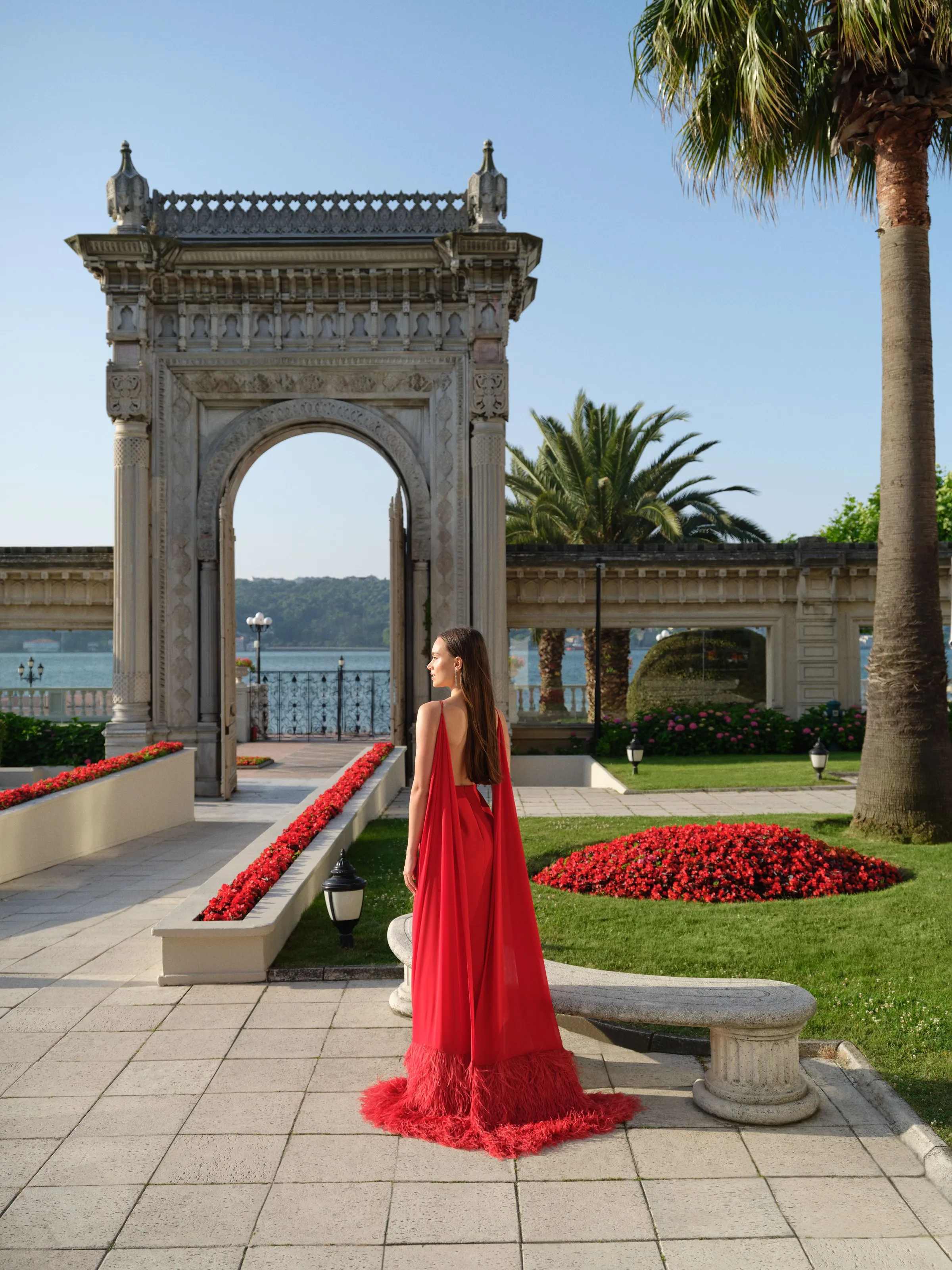 A woman in a red gown stands under the stone arch of the palace gate with the Bosphorus behind.