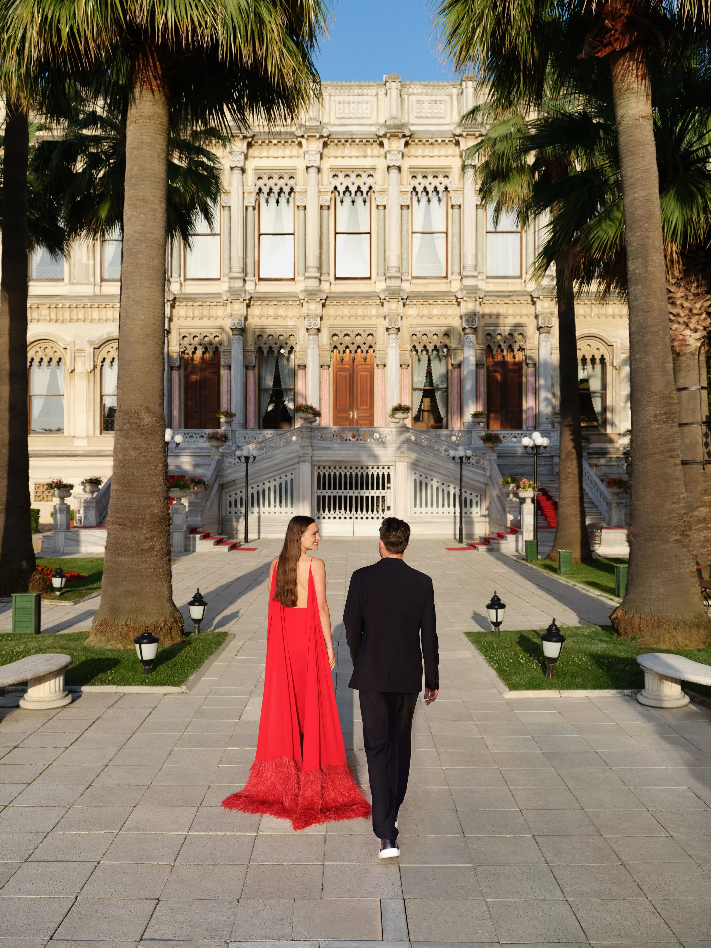 A couple in evening dress walk toward the palm-lined façade of Çırağan Palace Kempinski at dusk.