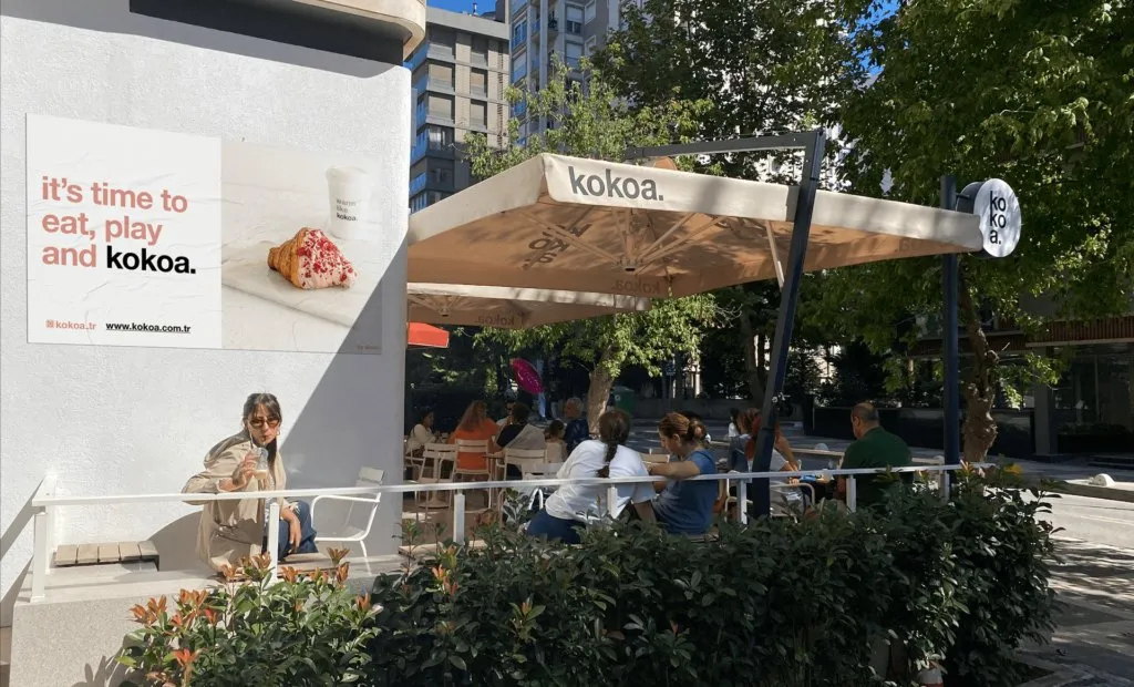 The café terrace under a kokoa parasol, with a poster reading 'it's time to eat, play and kokoa.'