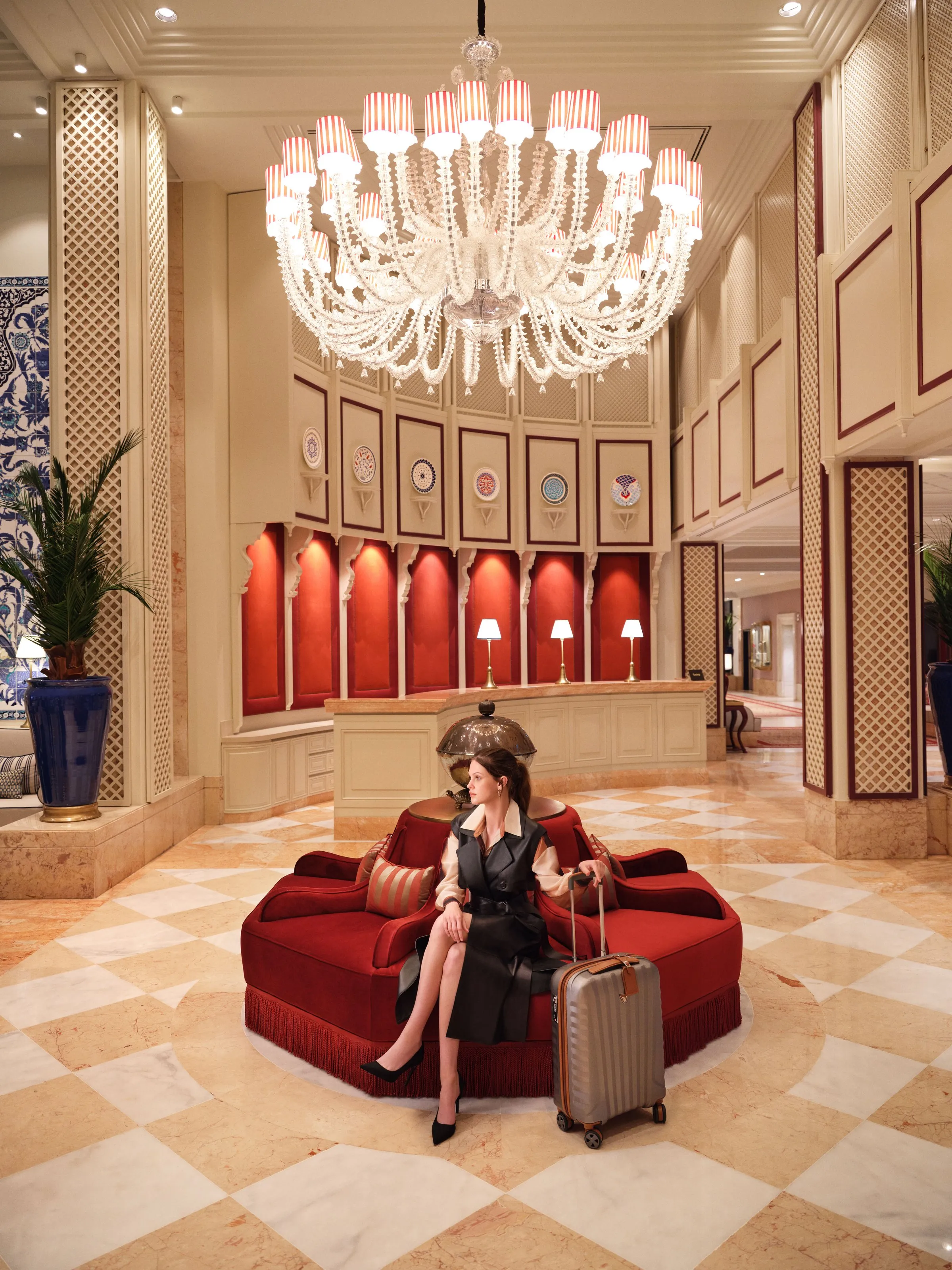 A traveller with a suitcase on a red circular sofa beneath a chandelier in the lobby.