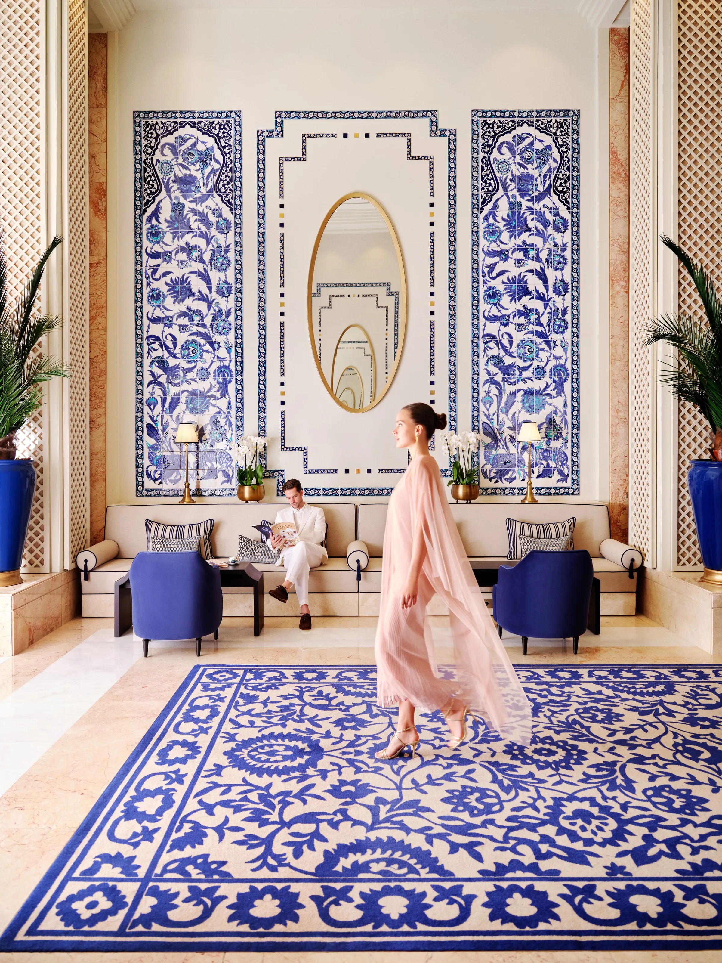 A guest in a pale gown crosses the lobby in front of blue Iznik-tiled walls and a patterned carpet.