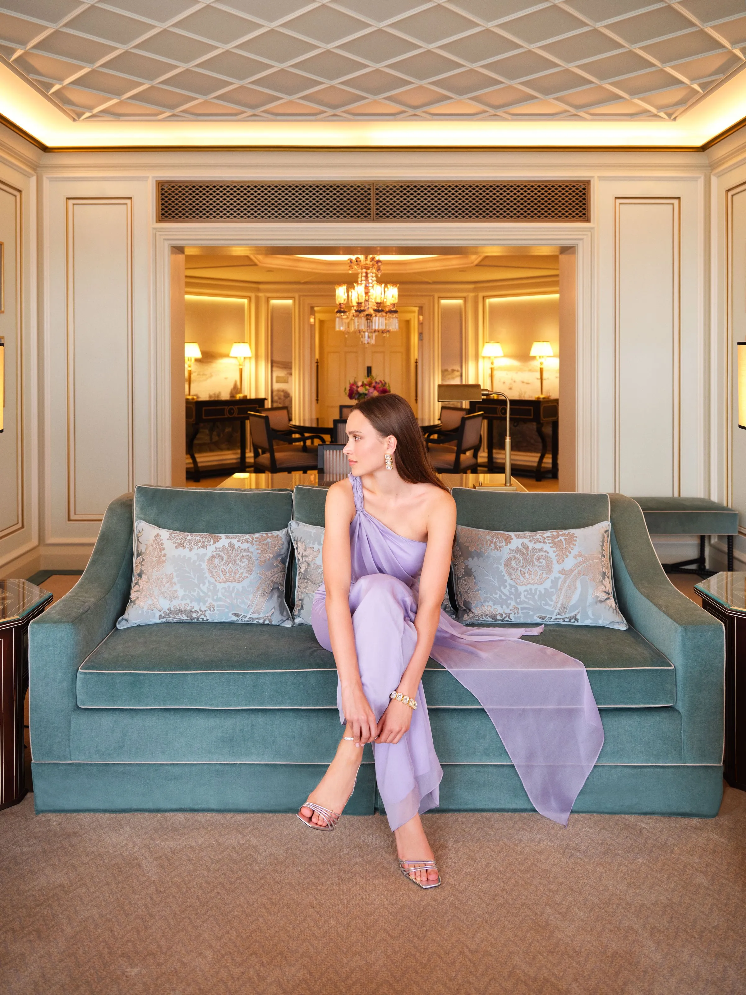 A guest seated on a turquoise sofa in a suite lounge with chandeliers and cream panelling.